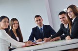 3 Steps to Use the Pygmalion Effect for Performance. Image shows five people in business attire sitting at a table, looking toward the camera. The group is made up of three women and two men.