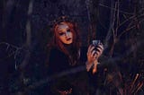 A pale red headed woman dressed in black holds a cup of a mysterious liquid in the middle of the forest