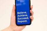 A hand holding a cell phone with the words believe, achieve, succeed, repeat.