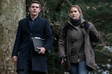 Two characters dressed for cold weather walk towards the camera in the woods. From left to right they are Colin Zabel played by Evan Peters and Mare Sheehan played by Kate Winslet.