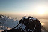 Four people standing on a rocky mountain topped with snow, more rocky peaks and the sunset in the background