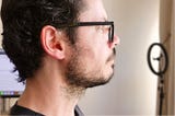 White man’s face, with glasses and beard, in profile, with emphasis on a pink plug in the ear.