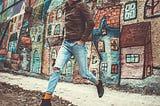 A stylish man in a casual outfit against a vibrant, colorful mural depicting whimsical houses. He seems to be walking on the air.