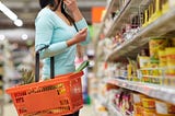 woman wearing teal sweater carrying shopping basket pauses in a supermarket aisle to consider the options