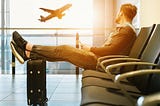 Man sitting in airport terminal with feet up on suitcase looking out the window as a plane takes off.