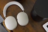 A pair of white Sonos Ace headphones on a wooden desk. A black speaker next to it and the corner of an iPad bottom right.