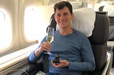 Nomadic Matt sitting in business class on a plane, holding up a glass of champagne