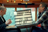 Julian Clary with his hand covering his mouth as he laughs, the table in the caravan destroyed
