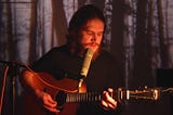 Bo Burnham, a bearded man, is sitting in a room playing the guitar and singing with trees projected onto the wall behind him