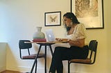 Manivillie sits by a small round wooden table typing on a silver Macbook. There is a small notebook in front of the Macbook