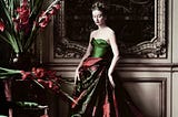 A model wearing a plaited up-do and a discreet wreath of red berries and green leaves poses in a green tafetta strapless bodice and a large skirt of dark green and berry red swagged at the waist. Gladioli in a vase match the berry red of the dress skirt and spill out above a green velvet seated chair. The model poses against a light brown panelled background with a dark chinosserie painting behind her. It’s all very country house ball.