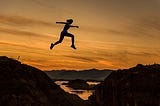 Young Woman In Sport Apparel Jumping Over A Cliff With Sunset In The Background.
