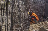 Man wearing hard hat and orange safety vest is tying iron construction rods together.