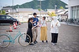 Photo of four people looking at a sheet of instructions. They are outside, on the plaza of a school, with mountains behind.