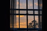 A view of the sky at dusk, with the silhouette of the window frame and sheer curtains in front.