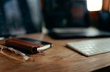Shallow depth of field photo with partial focus on a small notebook with a leather pen holder on the spine and an external keyboard, with a laptop in the background