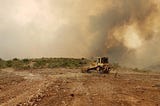 A bulldozer on a stretch of brown dirt, in front of a sky blotted out by smoke clouds.