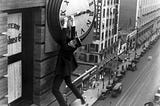 A black and white image of a silent movie. A man with specs and a flat, straw hat hangs off the hand of a clock on the side of a tall building in the city.