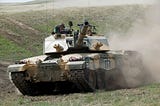 A Challenger 2 speeds down a green grassy hill in an exercise. The Challenger has sloped frontal armour on a massive boxy turret.