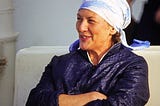 a woman with a blue jacket and light blue headscarf, smiling and looking confidently