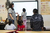 The image shows a classroom scene with a teacher and several young students. The teacher, standing on the left, is interacting with the students, some of whom are seated while others are standing near a large interactive whiteboard. One student is writing on the board, which displays the word “vest.” Another student is seated with their hand raised, indicating they may have a question or want to participate. The classroom has various educational posters on the walls, including one with basic key