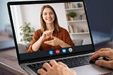 Video call on a laptop screen with a woman in a brown shirt using sign language. Books and plants in the background. Hands typing.