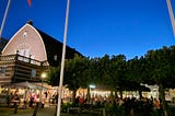 A historic, red brick building at twilight with a cozy beer garden next to it brimming with life against a cloudless sky