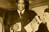 Smiling very tall man wearing a suit and carrying a newspaper. To his right and half cropped from the photograph is a woman smiling and looking upwards towards him