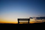A silhouette of an empty bench in a grassy field at sunset.