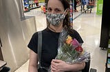 A masked Rebecca holding a bouquet of roses while sitting on her scooter in a grocery store