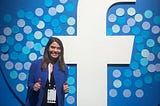 Laurie Keith stands in a blue jacket and black jeans in front of a giant Facebook logo with various blue dots.