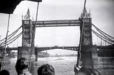 A black and white photograph depicts the Tower Bridge in London as seen from the perspective of individuals on a boat in the river.