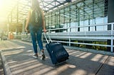 Une femme avec un sac à dos tire une valise en direction d’une gare de train ou un aéroport.