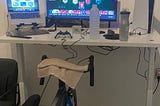 This photo shows a home bike training station setup. The bike is mounted on an indoor trainer stand, and there are two screens on the desk. A game is open on the large screen, while another application or program is running on the smaller one. On the desk, there are a water bottle, towel, and other equipment. A wall-mounted shelf holds decorative items, small plants, and framed photos. On the right side, there’s a large white device, likely a gaming console.