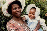 Black woman in a hat holding a small Black baby wearing a bonnet.
