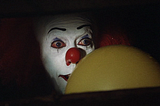 Tim Curry as the clown in Stephen King's It, holding a yellow ballon in a storm drain, looking overly friendly