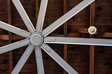 Nine bladed silver industrial fan against a wood ceiling.