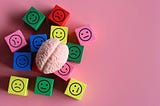 Quadradinhos de madeira com a silhueta de rostinhos com expressões felizes, tristes e neutras, em cima de um fundo rosa com um cérebro de plástico em cima dos quadradinhos.