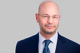 jason colodne wearing a blue jacket and a blue tie with glasses in front of a gray background