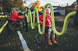 Duas crianças de pele branca, em cadeiras de roda, vestindo roupas de inverno. Brincam em um parque projetado com design inclusivo.