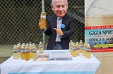 A man wearing a suit and a Netanyu mask points to a plastic bottle filled with yellow-colored liquid and labeled “Gaza Springs” as he stands behind a table with more bottles and some literature, next to a large poster showing an enlarged bottle with the label saying “Bottled at the source — GAZA SPRINGS — Complete Siege Sewage and Seawater — ‘A Taste of Genocide.’”