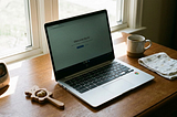 A laptop with a Chrome sticker, the screen says “Welcome back”. There is a baby rattle, coffee mug, baby bag and baby monitor on the desk.