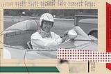 A black-and-white photo of a woman wearing a polka-dot helmet, with facing goggles around her neck, smiling from the driver’s seat of a racecar.