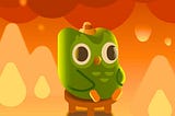 The green Duolingo owl sits among flames in a parody of the “this is fine” meme.