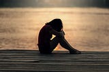 A young lady sitting at the docks with her face hidden on her forearms resting on her knees