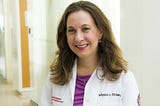 Dr. Allyson Ocean standing by window wearing a purple shirt and her white medical coat.