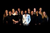 A multi-racial, multi-generational group of 13 people standing together, facing the camera. Some are smiling, some look smug.