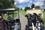 Photo of the back of two golf carts with 3 sets of golf clubs. The Carts are at the tee blocks of the par three number nine hole facing the green.