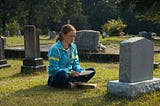 A still from the episode featuring Max, a young teenage girl with red hair pulled back in a ponytail and a blue windbreaker, sitting cross-legged in front of a headstone, holding a piece of paper in her hand.