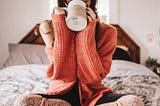 Woman in red knit sweater sitting on bed sipping out of a mug